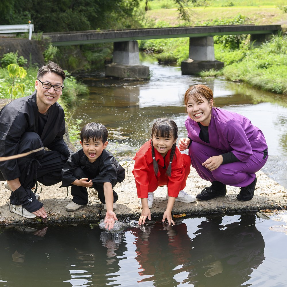 里山の川辺で自然に触れる忍者衣装の家族